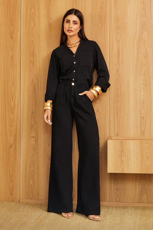 Mulher vestindo conjunto preto elegante, com camisa de mangas longas e calça de alfaiataria ampla, posando em ambiente minimalista com painel de madeira ao fundo.